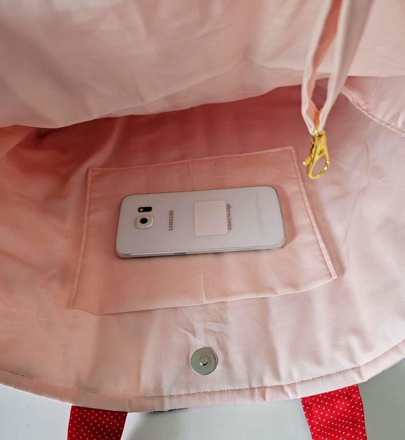 Puede incluir: Bolso tote rosa con asa de lunares rojos y un bolsillo. Un tel&eacute;fono Samsung Galaxy plateado descansa dentro del bolsillo. El bolso tiene un cierre magn&eacute;tico a presi&oacute;n y un cierre dorado.