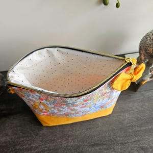 May include: A zippered pouch featuring a colorful floral and character print on the upper section and a solid mustard yellow base. The interior is lined with white fabric featuring small gold dots. A yellow fabric bow is attached to the side zipper.