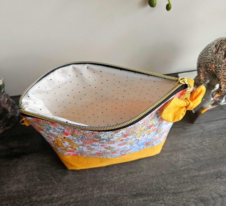 May include: A zippered pouch featuring a colorful floral and character print on the upper section and a solid mustard yellow base. The interior is lined with white fabric featuring small gold dots. A yellow fabric bow is attached to the side zipper.