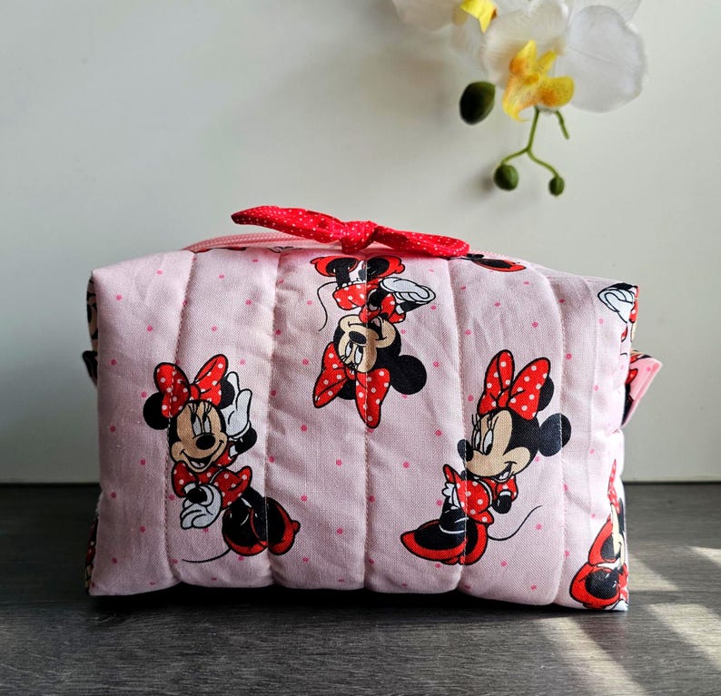 Puede incluir: Un neceser acolchado rosa con Minnie Mouse en varias poses. La bolsa tiene un lazo rojo de lunares y un cierre de cremallera. La bolsa mide aproximadamente 20 cm de largo y 13 cm de alto.
