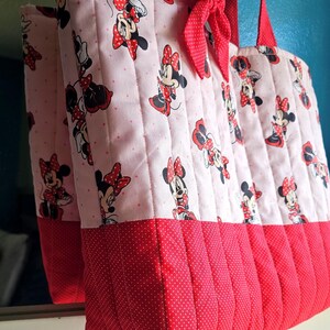 Puede incluir: Un bolso tote acolchado con un estampado de Minnie Mouse sobre un fondo rosa claro con lunares rojos. La parte inferior es roja con un dise&ntilde;o de lunares. El bolso tiene asas rojas y un lazo rojo.