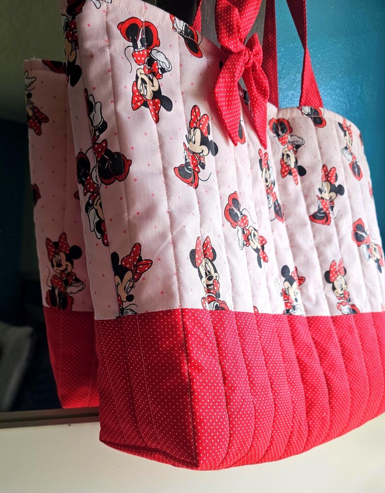 Puede incluir: Un bolso tote acolchado con un estampado de Minnie Mouse sobre un fondo rosa claro con lunares rojos. La parte inferior es roja con un dise&ntilde;o de lunares. El bolso tiene asas rojas y un lazo rojo.