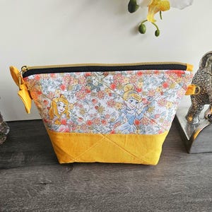 May include: A trapezoidal zippered pouch featuring a fabric print of two animated princesses amidst a floral pattern on a white background. The bottom section is solid mustard yellow fabric with quilted stitching, and the top has a black zipper.