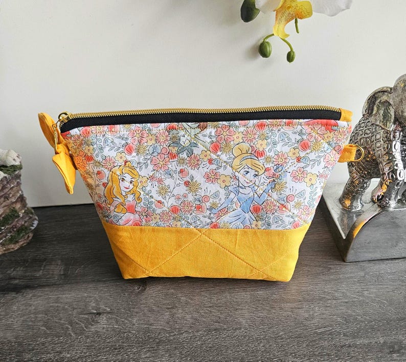 May include: A trapezoidal zippered pouch featuring a fabric print of two animated princesses amidst a floral pattern on a white background. The bottom section is solid mustard yellow fabric with quilted stitching, and the top has a black zipper.