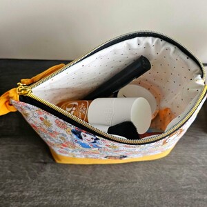 May include: A fabric zipper pouch featuring a colorful Snow White and floral print on a white background with a solid yellow base. The interior is lined with white fabric featuring small black dots and contains a black makeup brush and a white bottle.