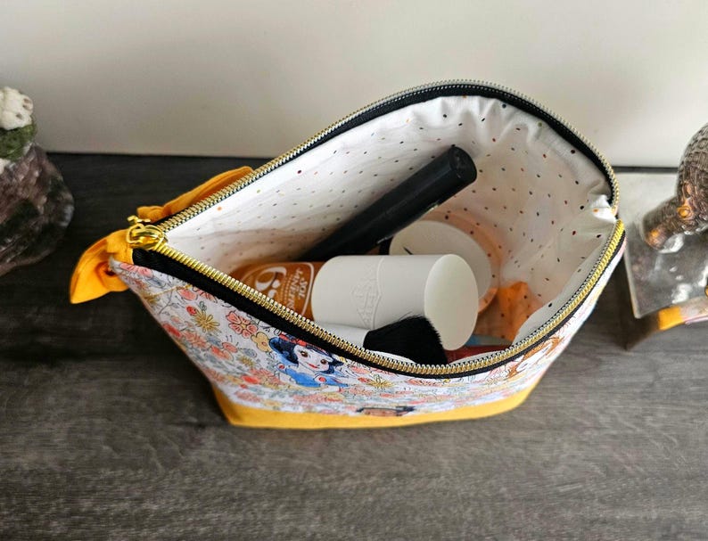 May include: A fabric zipper pouch featuring a colorful Snow White and floral print on a white background with a solid yellow base. The interior is lined with white fabric featuring small black dots and contains a black makeup brush and a white bottle.
