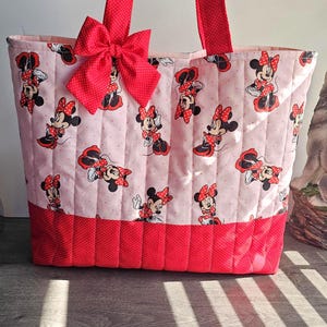 Puede incluir: Bolso tote acolchado con un estampado de Minnie Mouse sobre un fondo rosa claro. El bolso tiene detalles de lunares rojos, un gran lazo rojo y asas rojas. Dise&ntilde;ado para transportar objetos.
