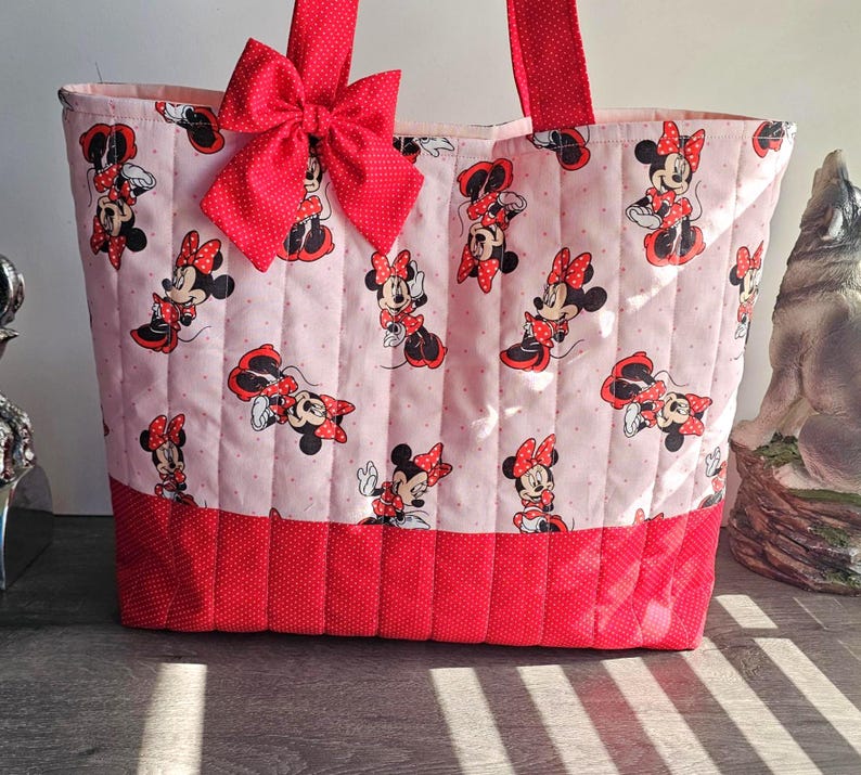 Puede incluir: Bolso tote acolchado con un estampado de Minnie Mouse sobre un fondo rosa claro. El bolso tiene detalles de lunares rojos, un gran lazo rojo y asas rojas. Dise&ntilde;ado para transportar objetos.