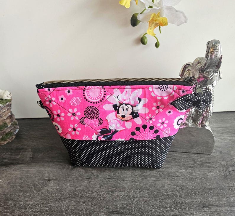 Peut inclure: Une pochette en tissu matelass&eacute; avec un fond rose vif, un imprim&eacute; Minnie Mouse, des motifs floraux blancs et des pois noirs. La partie inf&eacute;rieure est noire &agrave; pois blancs et elle est dot&eacute;e d'une fermeture &eacute;clair en m&eacute;tal.