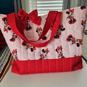 Puede incluir: Bolso tote acolchado con fondo rosa claro y estampado de Minnie Mouse. Cuenta con detalles de lunares rojos, un lazo rojo y correas rojas. El bolso tiene una base rectangular y una amplia abertura, ideal para llevar cosas.