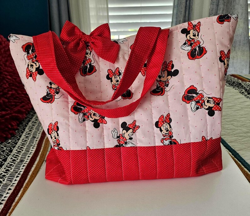 Puede incluir: Bolso tote acolchado con fondo rosa claro y estampado de Minnie Mouse. Cuenta con detalles de lunares rojos, un lazo rojo y correas rojas. El bolso tiene una base rectangular y una amplia abertura, ideal para llevar cosas.