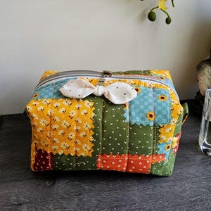 May include: A patchwork cosmetic bag with a bow-shaped zipper pull. The bag features a variety of fabrics in yellow, green, blue, and red, with floral and polka dot patterns. The bag is rectangular and has a silver zipper.