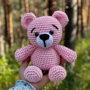 May include: A pink crocheted teddy bear with black eyes and a white nose. The bear is sitting on a hand.