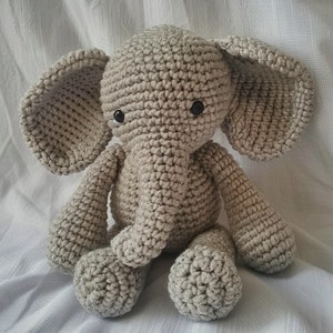 May include: A crocheted gray elephant toy with black eyes. The elephant is sitting on a white surface.
