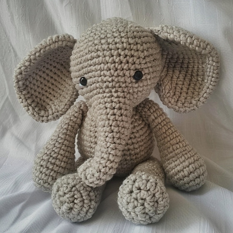 May include: A crocheted gray elephant toy with black eyes. The elephant is sitting on a white surface.