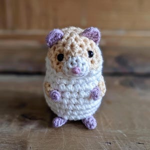 May include: A small crocheted hamster toy with white fur, light brown accents, and purple ears and feet.