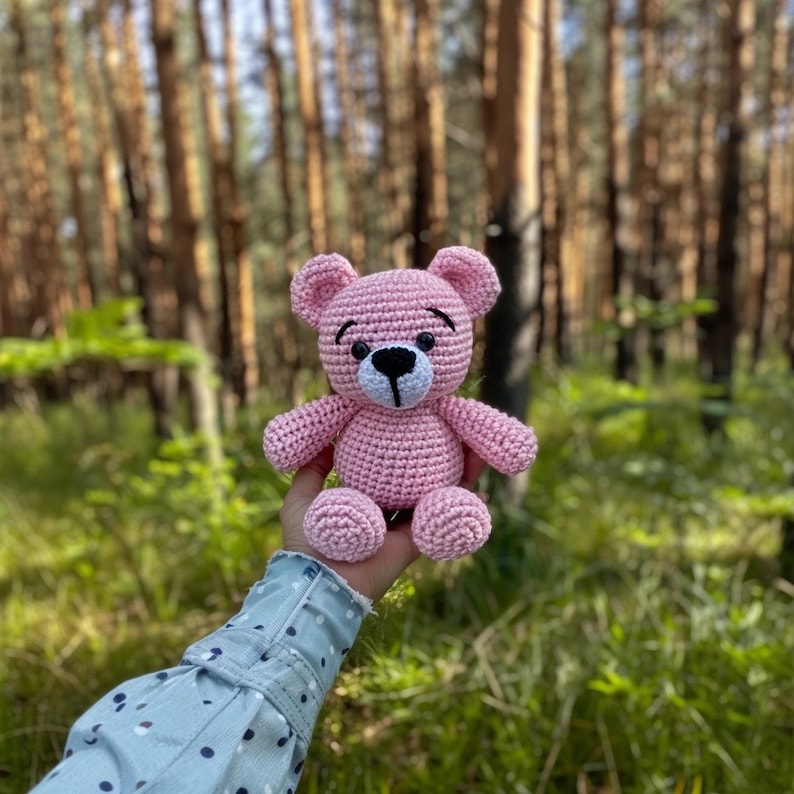 May include: A pink crocheted teddy bear with black eyes and a white nose. The bear is sitting in the palm of a hand.