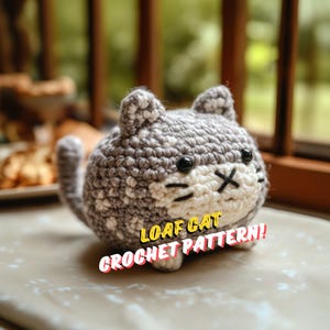 May include: A crocheted cat toy in shades of gray and white, with black eyes and a stitched mouth. The cat is in a loaf position. The text "LOAF CAT CROCHET PATTERN!" is displayed in a yellow and red banner.