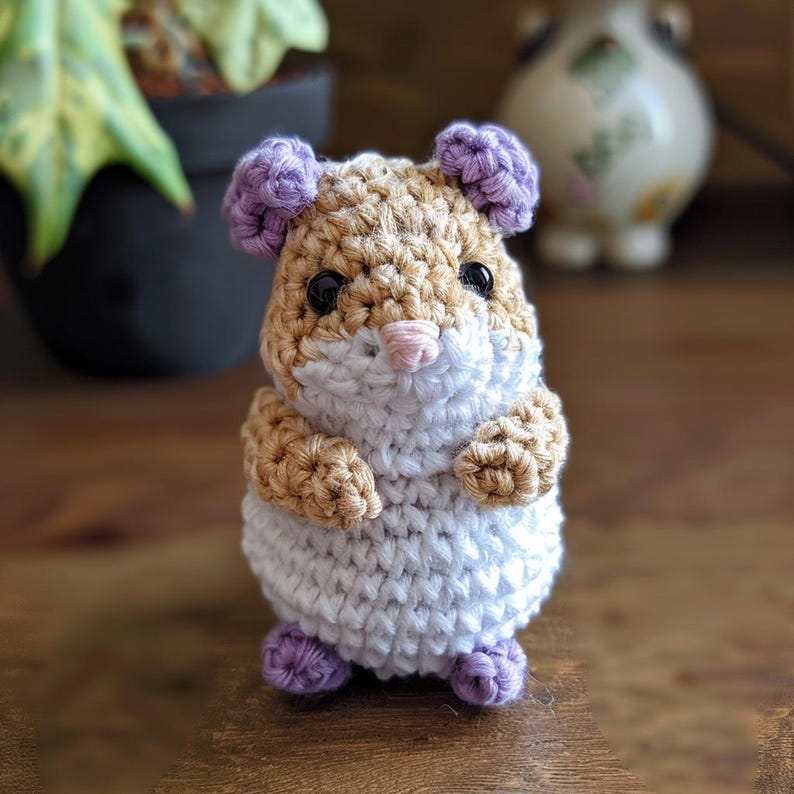 May include: A crocheted hamster toy with a white body, brown and tan fur on its head and arms, and purple ears and feet.