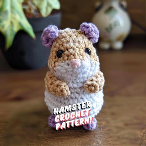 Puede incluir: Un juguete de crochet en forma de hámster con pelaje marrón y blanco, orejas moradas y ojos negros. El juguete está sentado sobre una superficie de madera y tiene el texto "HAMSTER CROCHET PATTERN!" en su vientre.