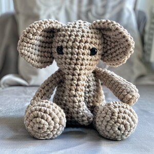 May include: A crocheted elephant toy with a beige color and black eyes. The elephant is sitting with its legs crossed and its trunk pointing straight ahead.