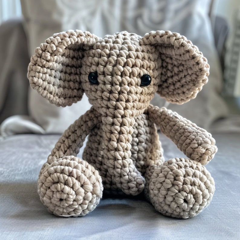 May include: A crocheted elephant toy with a beige color and black eyes. The elephant is sitting with its legs crossed and its trunk pointing straight ahead.