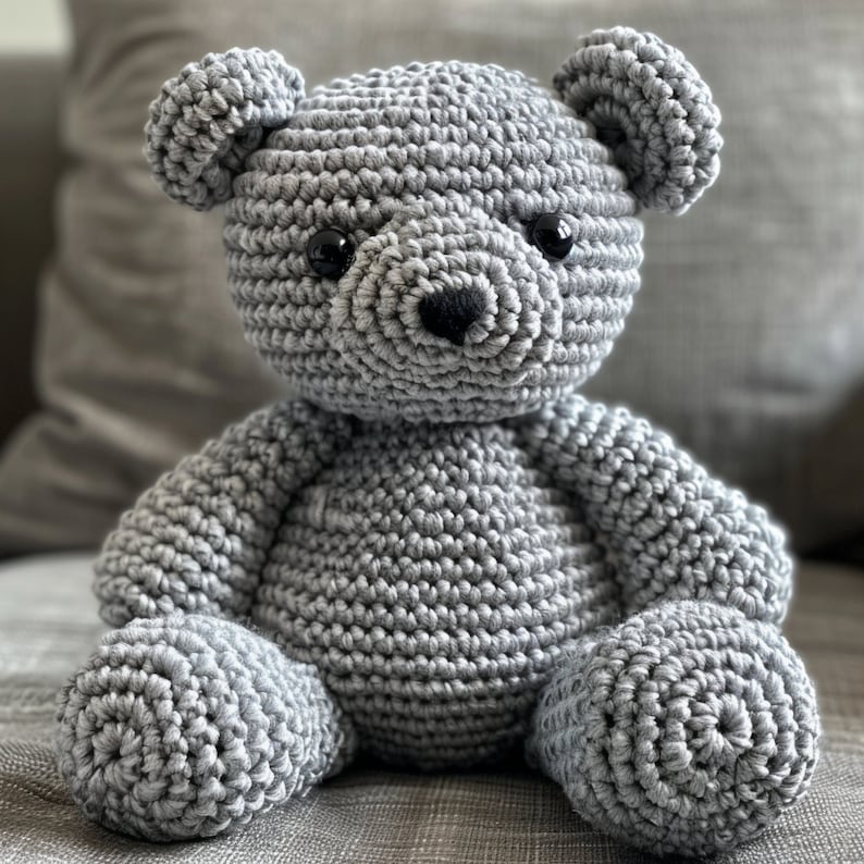 May include: A gray crocheted teddy bear with black eyes and a black nose. The bear is sitting on a gray couch.