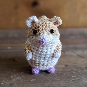 May include: A crocheted hamster toy with brown and white fur, black eyes, and a purple nose. The toy is standing on a wooden surface.