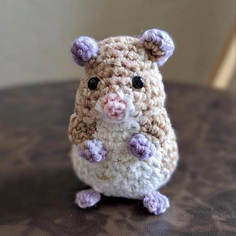 May include: A small crocheted hamster toy with brown and white fur, purple ears, and purple feet. The hamster has black eyes and a pink nose.