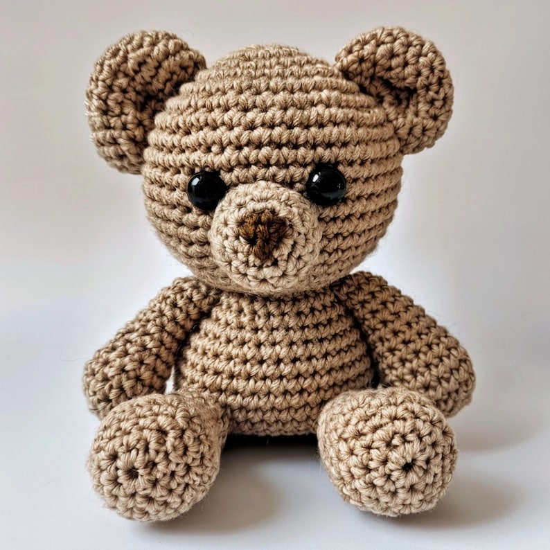 May include: A crocheted brown teddy bear with black eyes and a brown nose. The bear is sitting with its legs crossed.