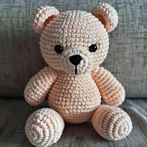 May include: A crocheted teddy bear with a light peach color. The bear has black eyes and a black nose. The bear is sitting on a textured surface.