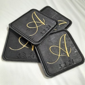 May include: Four black square cork coasters with a gold embroidered letter A and the date 3.2.24.