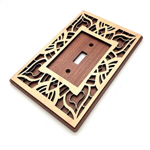 May include: Wooden light switch cover with a decorative, laser-cut design. The cover features a rectangular shape with a dark brown center and a light brown, patterned border. The switch opening is a standard toggle style.