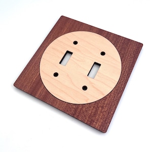 May include: A wooden double light switch cover. The cover has a square, dark brown frame with a circular, light-coloured centre. The centre has two rectangular openings for the switches and four screw holes.