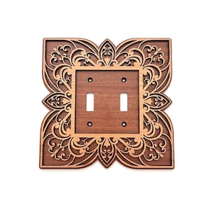 May include: A decorative wooden double light switch cover with an intricate floral design. The cover is made of two shades of brown wood, with a darker frame and a lighter, detailed floral pattern. The switch openings are rectangular.