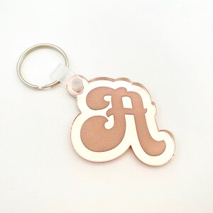Könnte beinhalten: Ein roségoldfarbener Schlüsselanhänger mit dem Buchstaben "A" und weißer Umrandung. Der Schlüsselanhänger hat einen silbernen Metallring und einen transparenten Kunststoffverbinder. Der Buchstabe ist in einem Retro-Schriftstil gehalten.
