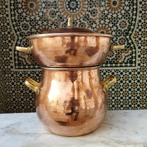 May include: Two-piece copper cookware set with brass handles. The set includes a pot and a lid, stacked on top of a larger pot. The copper has a hammered texture, and the handles are a bright gold color. The background features a mosaic tile design.