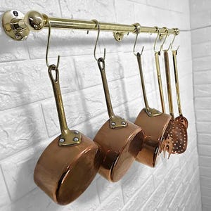 May include: A set of copper-bottomed saucepans and kitchen utensils suspended from a brass rail against a white brick wall. The pans have brass handles. The utensils include a slotted spoon and a fork.