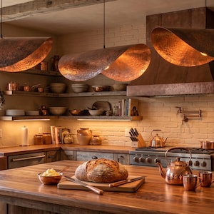 May include: Three large, curved, hammered copper pendant lights hang above a wooden kitchen island. The lights feature a textured, metallic finish that reflects warm light downward onto the kitchen workspace and the loaf of bread below.