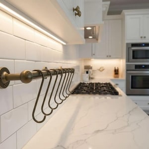 Peut inclure: Cuisine moderne avec des meubles blancs, un plan de travail en marbre et un four en acier inoxydable. Un porte-ustensiles en laiton avec des crochets est monté sur le mur carrelé blanc. Design épuré.