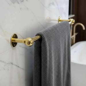 Peut inclure: Un porte-serviettes doré avec une serviette grise suspendue. Le porte-serviettes est fixé sur un mur en marbre blanc. Le porte-serviettes a un design de boule ronde aux extrémités.
