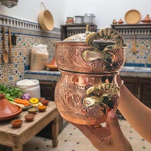 Pode incluir: Uma panela de cozinha de cobre de duas peças com padrões florais e de folhas ornamentados. A panela tem alças e uma tampa de latão. A panela é segurada em frente a uma cozinha com uma mesa de especiarias e uma panela tagine.