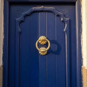 Peut inclure: Gros plan sur une porte en bois bleue avec un heurtoir doré décoratif. La porte a des panneaux verticaux et un sommet en arc. Le heurtoir est une poignée en forme d'anneau avec une base florale.