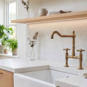 Peut inclure: Un robinet de cuisine de style pont avec une finition en laiton patiné, doté de deux poignées en croix et d'un bec haut. Le robinet est monté sur un plan de travail en pierre blanche à côté d'un évier timbre d'office et d'une petite assiette.