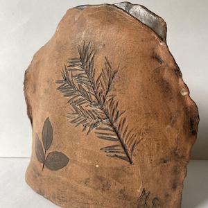 Könnte beinhalten: Ein handgefertigtes, braunes Keramikstück mit einer natürlichen, unregelmäßigen Form. Die Oberfläche zeigt geprägte botanische Designs, darunter ein detaillierter Farn und Blätter. Das Stück hat eine raue Textur und eine dunkelbraune Farbe, mit einer Signatur in der unteren rechten Ecke.