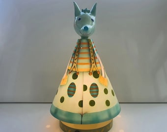 Puede incluir: Lámpara decorativa con forma de animal estilizado y orejas puntiagudas. El cuerpo cónico presenta lunares verdes claros y blancos, acentuados con rayas turquesas y naranjas. La base es circular y tiene un diseño decorativo.