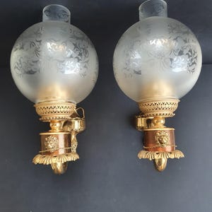 Könnte beinhalten: Zwei antike Wandleuchten mit Kugeln aus mattiertem Glas mit einem geätzten floralen Design. Die Wandleuchten haben Messing- und Kupferakzente, darunter ein dekorativer Löwenkopf. Auf dunklem Hintergrund montiert.