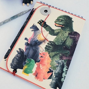 May include: A black Godzilla figurine with purple dorsal fins stands near a spiral-bound notebook. The notebook features a collage of colorful monster illustrations. A pencil rests beside the notebook.