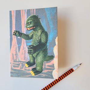 May include: A green monster figurine with yellow claws and teeth stands on a postcard. The postcard features a colorful cave scene. A pen with orange and black stripes is in the foreground.