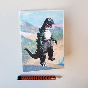 May include: A notebook featuring a Godzilla illustration with a mountain backdrop. The dinosaur is black, green, and blue. An orange and black striped pen rests below the notebook.